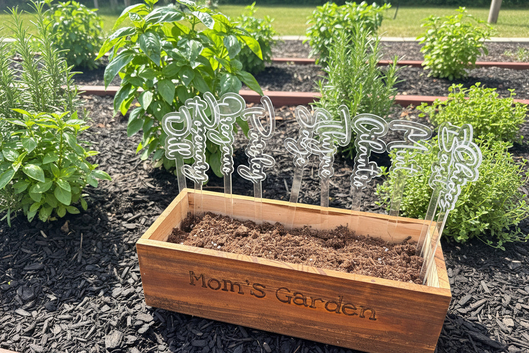 clear acrylic herb garden stakes with herb names placed in a wooden planter box surrounded by fresh herbs in an outdoor garden