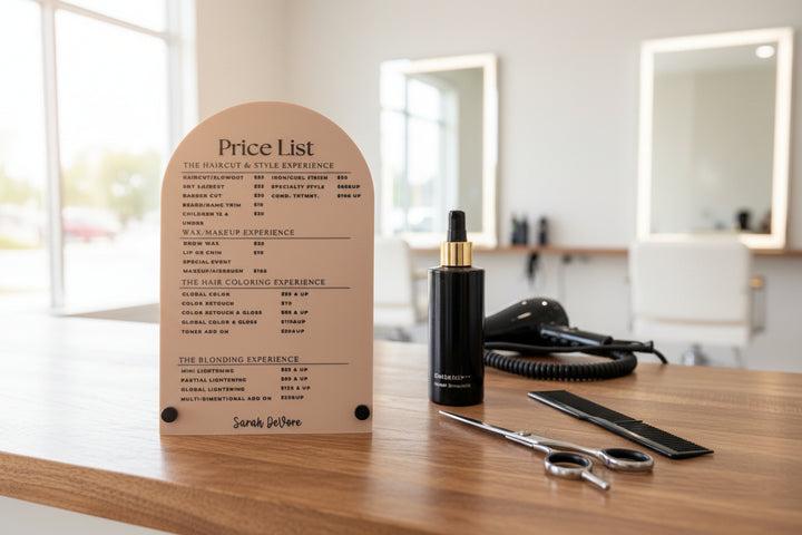 Arched frosted acrylic price list for a hair salon, featuring black text and a wooden base, displayed on a salon station with styling tools.