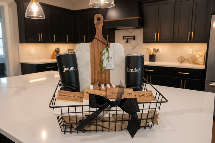 Complete Signature Plus gift basket on a white marble island, featuring a personalized marble and acacia board, black custom tumblers, and engraved coasters in a black wire basket.