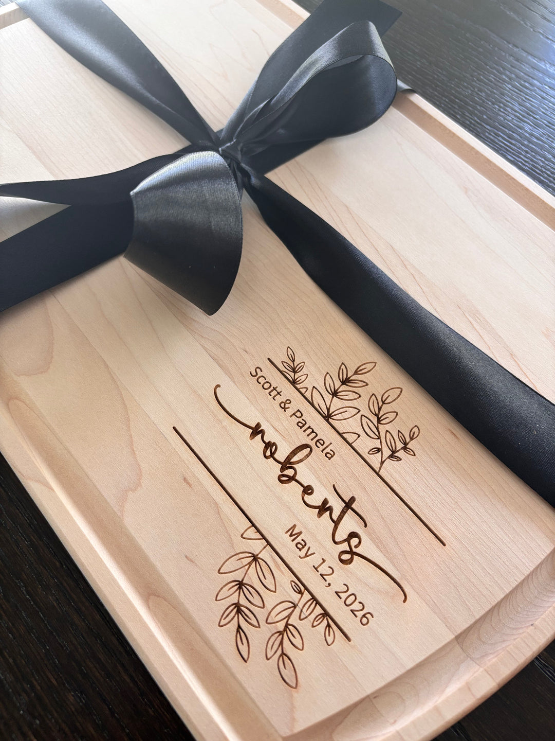 Large arched maple cutting board with custom laser-engraved wedding design, featuring names and wedding date, tied with a black ribbon.