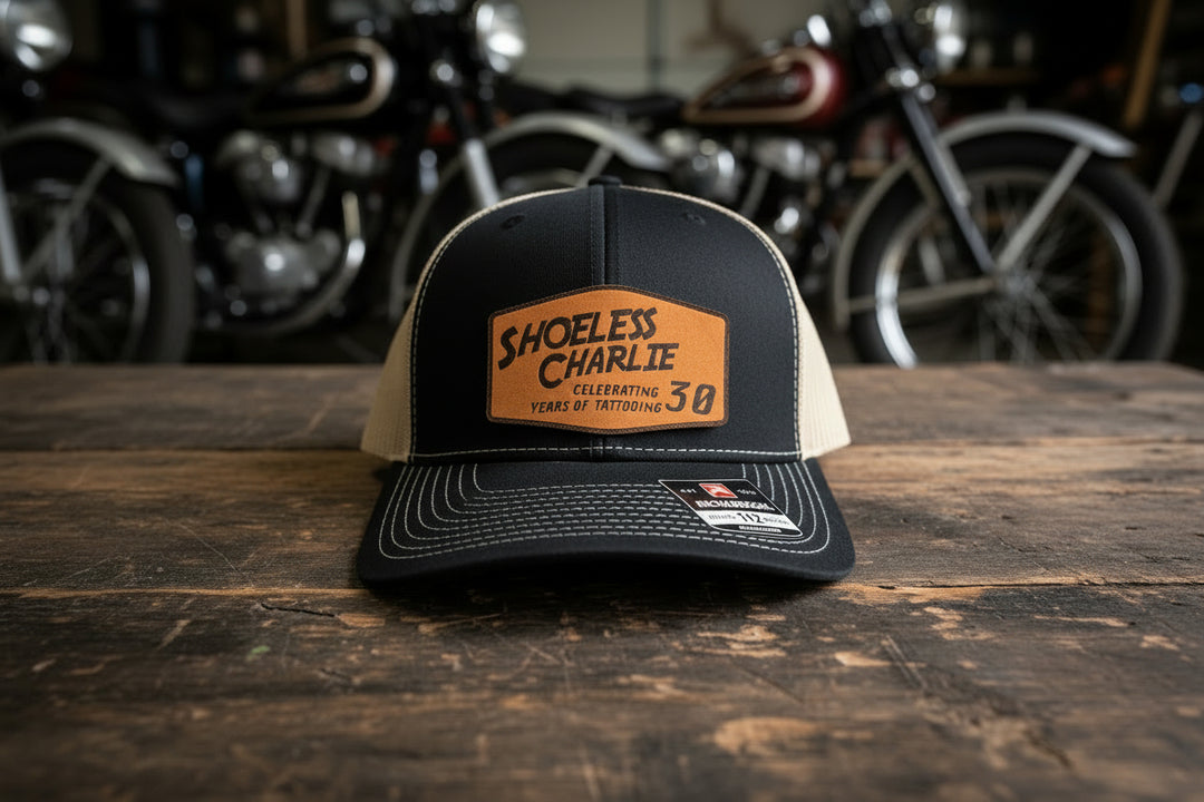 Shoeless Charlie 30th Anniversary Richardson 112 snapback hat with engraved leather patch displayed on wood table in motorcycle workshop.