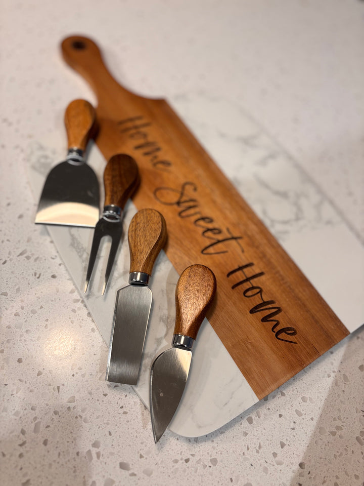 Set of cheese knives with wooden handles beside personalized marble and acacia charcuterie board engraved Home Sweet Home
